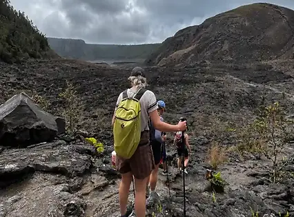 A hiker with a lime green backpack navigates across a rugged, rocky black volcanic lava field under a cloudy sky.
