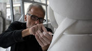 This image portrays an artist, whom we'll refer to as Dionisio Cimarelli, immersed in the intricate process of sculpting. He is attentively shaping a white sculpture with a delicate tool, indicating a moment of precise craftsmanship. The studio is bathed in natural light, enhancing the creative aura of the space. The artist's expression is one of deep concentration, reflecting his connection to the art and the skillful execution of his work