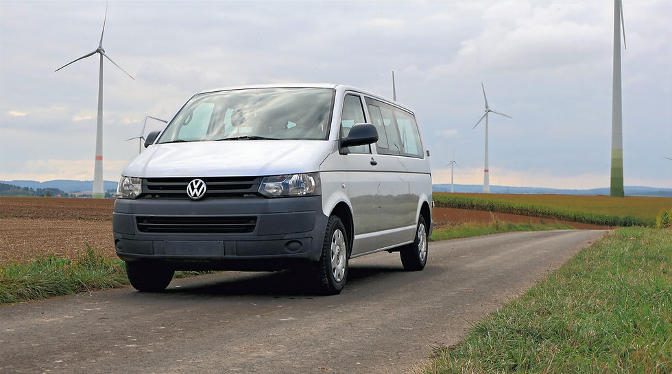 Silberner Volkswagen-Transporter fährt auf einer Straße mit Windkrafträdern und Feldern.