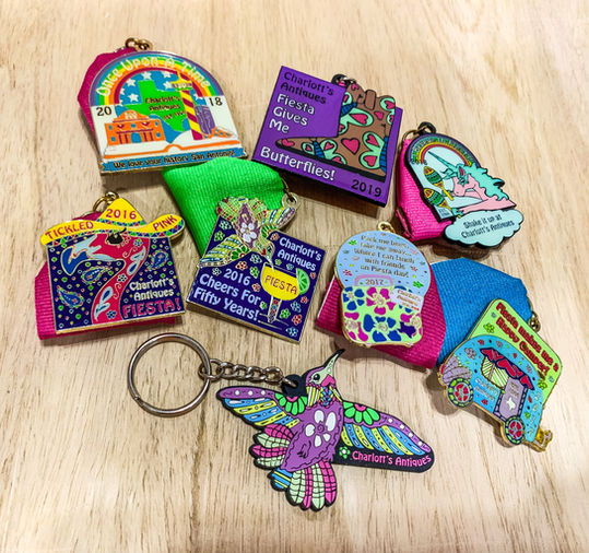 A group of 7 brightly colored Fiesta pins and a hummingbird keychain with Charlott's Antiques branding on them.