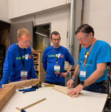Two members of COMO+Alt+Del standing next to tool tech David Fowler while he draws on a piece of wood