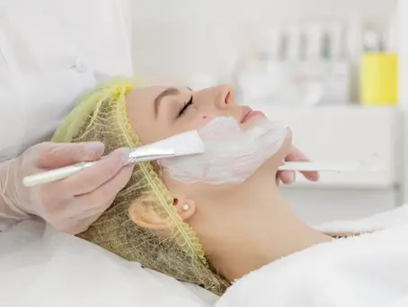 Profissional de estética aplicando máscara facial com pincel durante tratamento de pele em ambiente clínico.