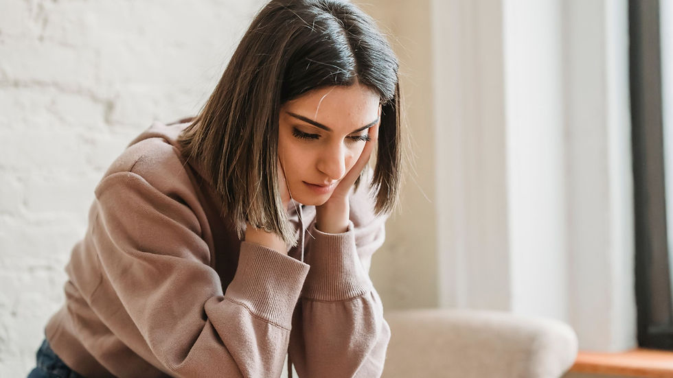 Woman in a beige sweater leans forward, head in hands, appearing thoughtful. Bright room with a brick wall and window in the background.
