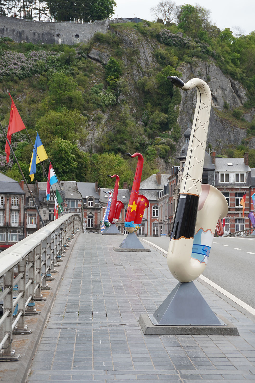 Colourful saxophone sculptures line a bridge adorned with international flags on the Charles De Gaulle Bridge in Dinant. Background features lush greenery and cliffs. No text visible.