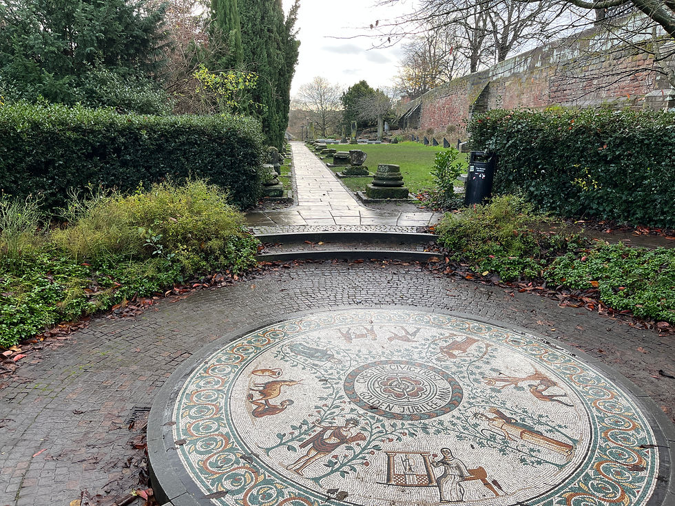 Mosaic with intricate patterns on a garden path, surrounded by lush greenery and a brick wall in a serene outdoor setting.