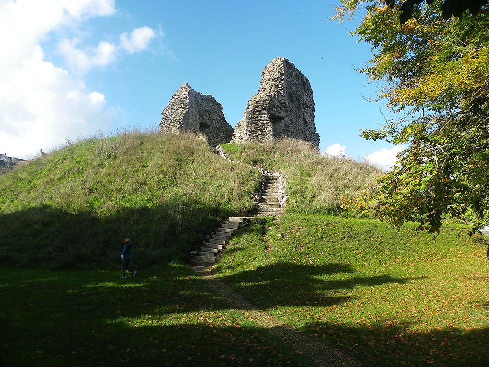 Ruins atop a grassy hill under a blue sky; shadowy foreground with a person walking near stone steps; trees partially frame the scene.