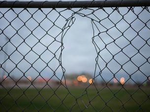 A chain-link fence with a torn opening, warm lights blurred in the distance beyond it.