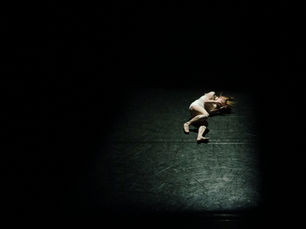 A figure curled small in a pool of light on a vast dark stage.