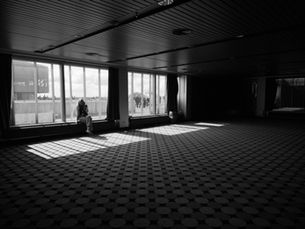 A solitary figure seated in a vast empty room, light falling through institutional windows.