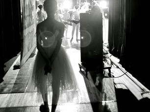A ballerina stands in shadow at the edge of a stage, watching a brightly lit group of dancers further down the corridor.
