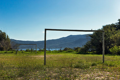Two concrete goalposts on a grass field beside a lake with forested hills, O Jogo, Leandro Martins