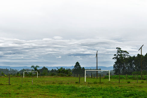 Two white goalposts on a vast green field with palm trees and layered clouds, O Jogo, Leandro Martins