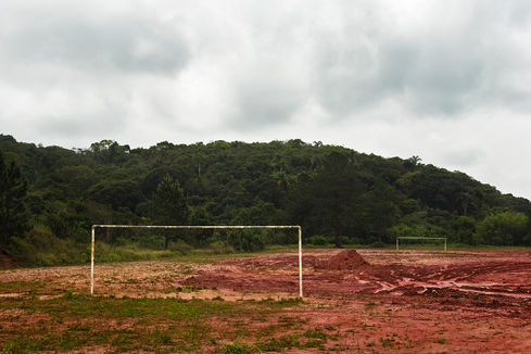 White goalpost on a red mud field with dense Atlantic forest hillside, O Jogo, Leandro Martins