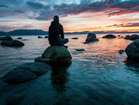 a man is sitting cross legs on a rock in a water body looking out at the sunset