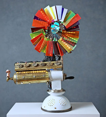 Kinetic sculpture with radial array of colored Plexiglas panels mounted above a brass canister and enamel grinder on a white strainer base