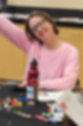 Smiling woman wearing a pink sweater and glasses. She is sitting at a table covered with legos, paper and a large water bottle