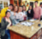 Photo of 7 friends standing around a kitchen counter and smiling at the camera while holding a Bundle board game.