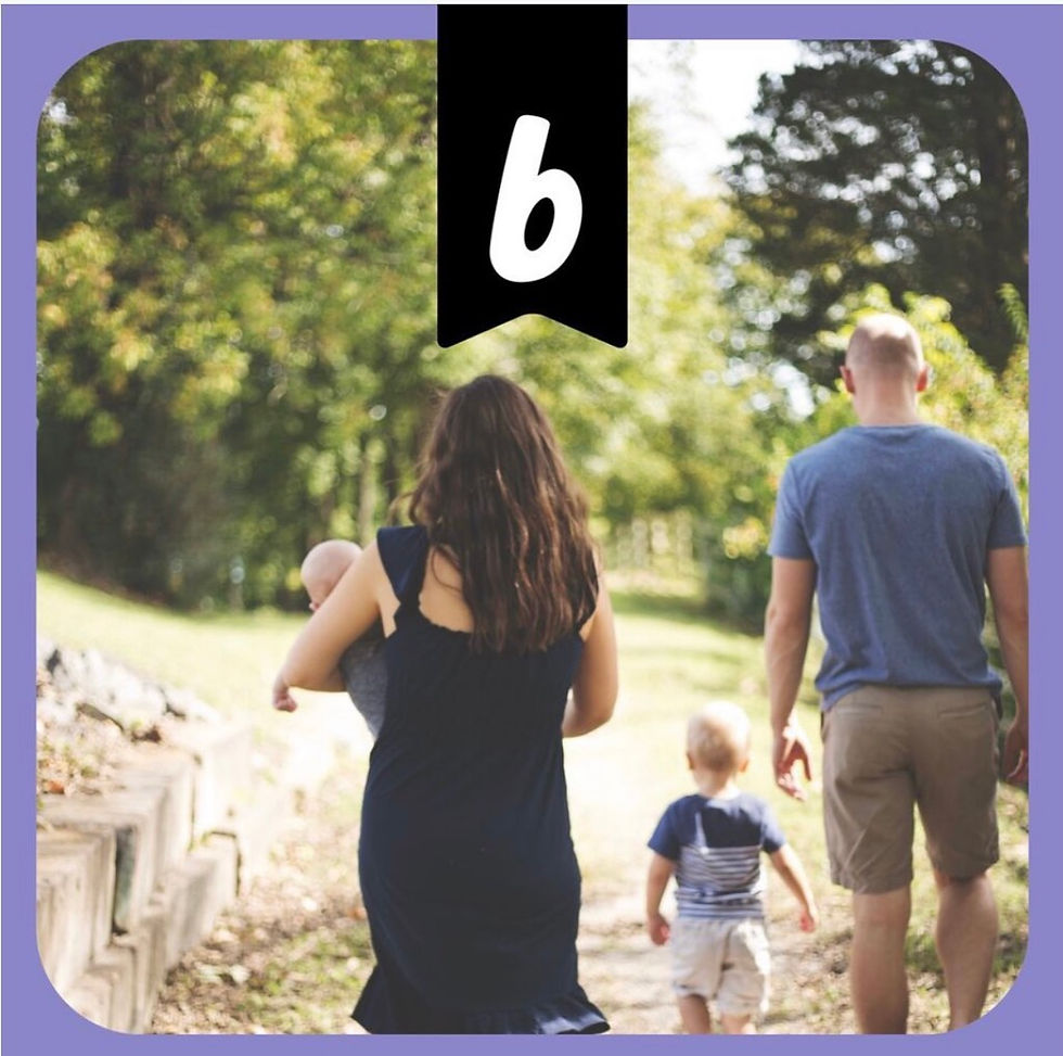 A family of four taking a walk through the woods. They are facing away from the camera. There is a mother in a black dress holding a baby. The father is walking a few steps ahead with a toddler.