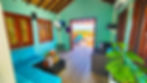 Cozy turquoise room with a blue sofa and TV, opening to a sunlit deck with a person in a hot tub. Bright, relaxing tropical vibe. Amber Cove Hillside Cabana