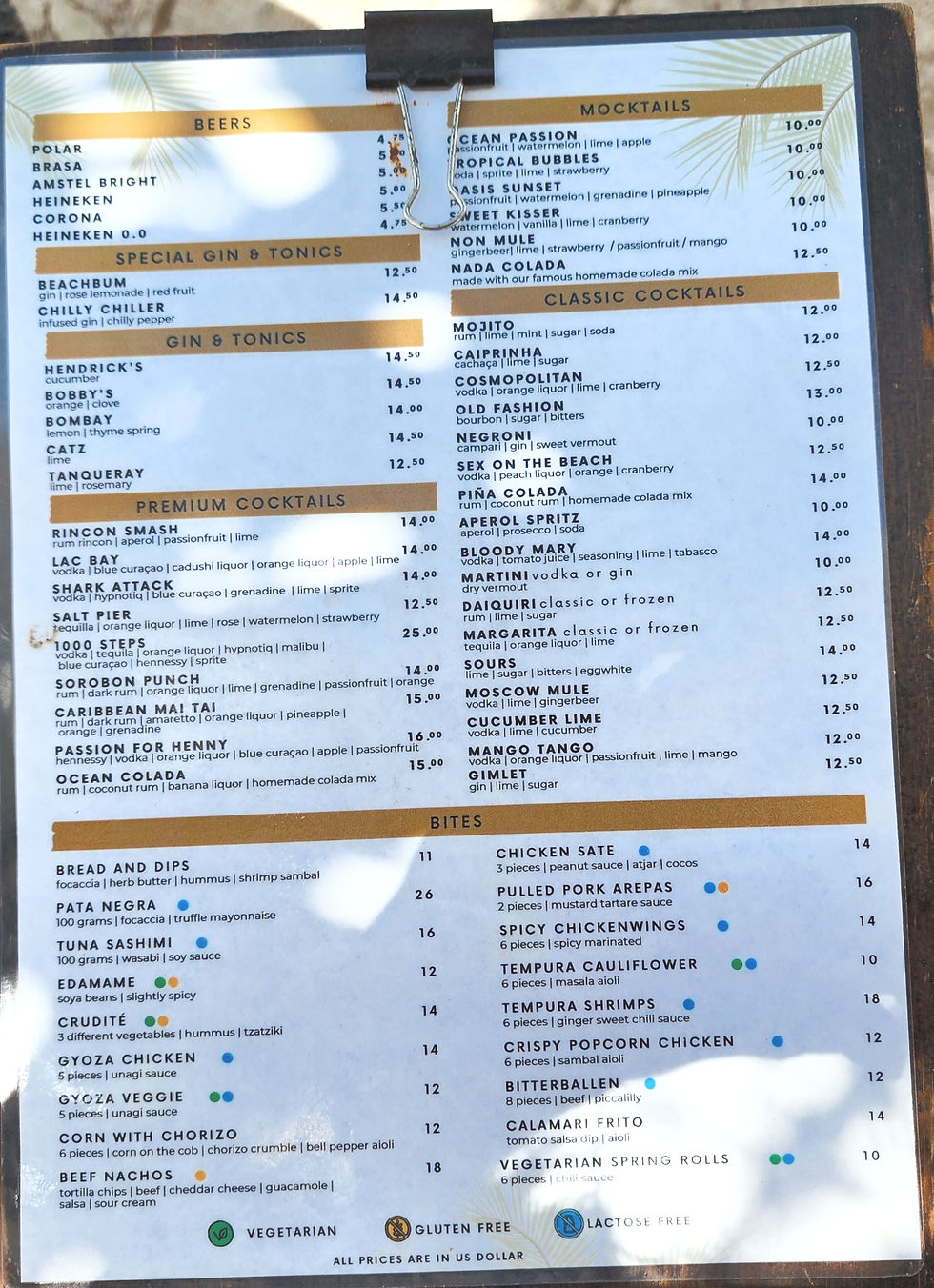 Beach bar menu board clipped with beers, mocktails, cocktails, and bites, clipped to a dark frame in bright sunlight. Ocean Oasis Bonaire