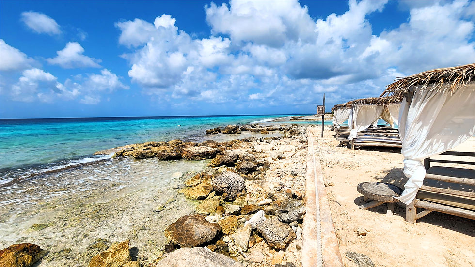 Sunny tropical beach with turquoise water, rocky shore, and thatched cabanas with white curtains under a bright cloudy sky
