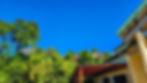Yellow house with green roof under a clear blue sky, surrounded by lush greenery and trees. Red patio cover in the foreground.