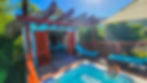 Child plays in a small pool on a sunny patio with tropical greenery. A cottage with open wooden doors and blue lounge chairs sets a relaxing mood.