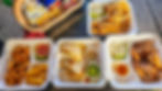 Takeout meals featuring onion rings, quesadillas, fries, and fried fish in foam trays on a wooden table, with a basket of chips and biscuits. Sky Bar Amber Cove Food