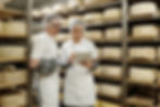 Two workers wearing hairnets and while lab coats inspecting cheese wheels with tablet in storage room. Shelves filled with aging cheese wheels surround individuals
