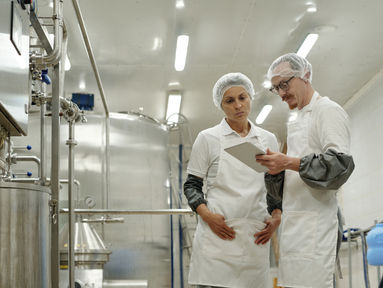 Kitchen staff in white uniforms and hairnets reviewing information on tablet at work. Industrial kitchen environment with large metal equipment and bright lighting