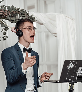 Ein junger DJ mit Brille, Kopfhörern und einem blauen Anzug mit Fliege steht vor einem Laptop. Er ist mitten in einer Ansage oder Moderation und gestikuliert mit der Hand. Im Hintergrund sind weiße Vorhänge und eine üppige Blumendekoration mit rosa und weißen Blumen zu sehen.