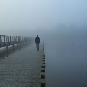 A person walks alone on a foggy pier over calm water. The setting is misty and tranquil, with grey tones which symbolizes the man's contemplation of therapy.