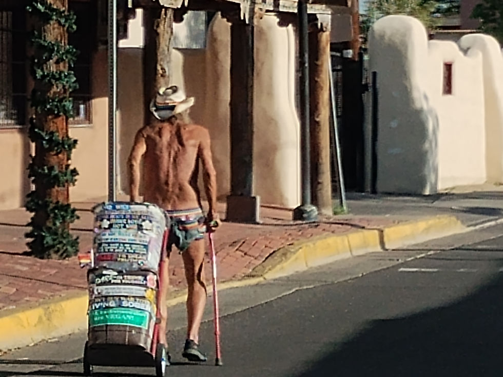 Man named Don with a hat and cane walks on a sunny street, pulling a cart covered in colorful stickers. Adobe buildings line the sidewalk.