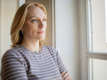 Mulher adulta loira olhando pela janela pensativa, representando a reflexão e a busca por compreensão no diagnóstico tardio do autismo em mulheres.