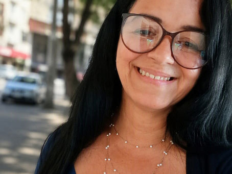 Mulher sorrindo de Cabelos longos lisos pretos de óculos na rua atrás tem uma árvore e um carro e o comercio local de saquarema rj.