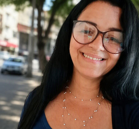 Mulher sorrindo de Cabelos longos lisos pretos de óculos na rua atrás tem uma árvore e um carro e o comercio local de saquarema rj.