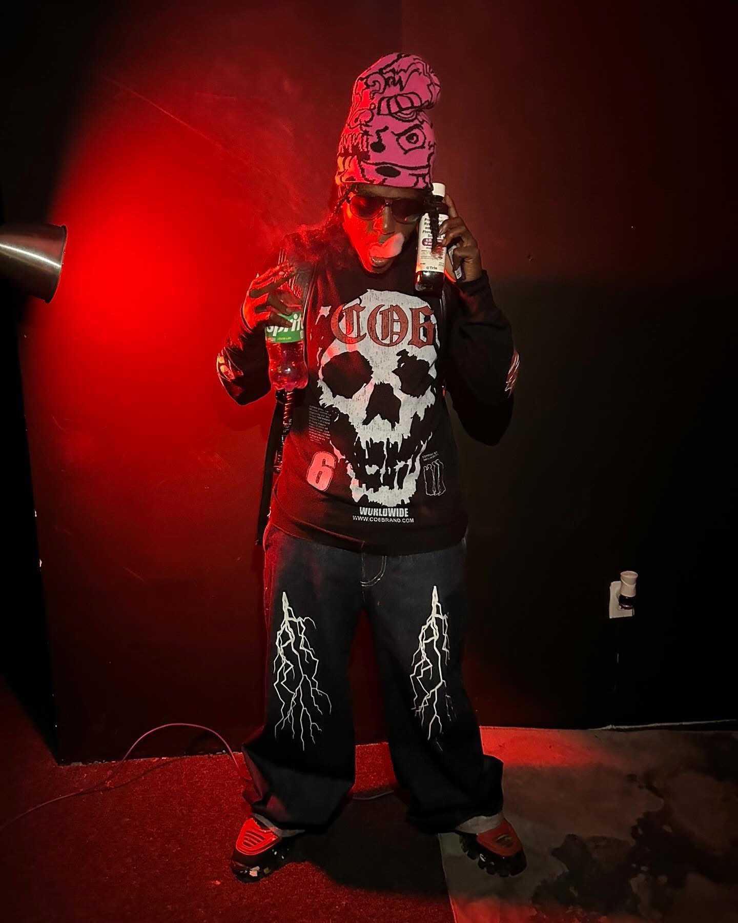 Trae Da Kidd de pie bajo una luz roja, con gorro rosa y jeans con estampado de rayos