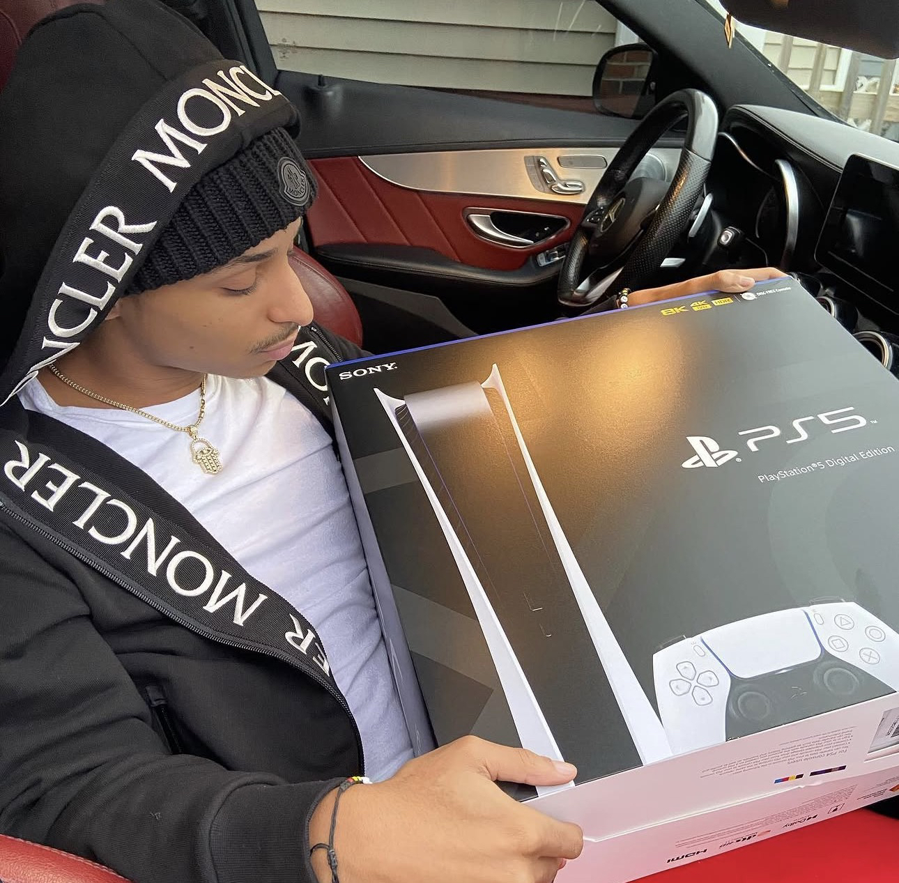 Cheeseaholic con un gorro negro, sosteniendo la caja de una PlayStation 5 dentro de un auto