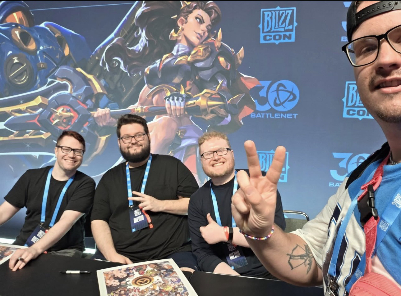 Tres hombres sentados a una mesa frente al arte de Overwatch y el logotipo de BlizzCon, con otro hombre haciendo la señal de paz en primer plano