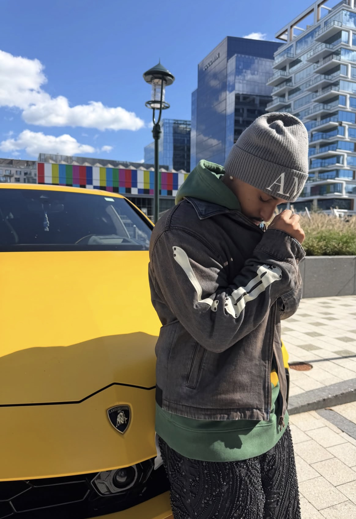 Cheeseaholic con un gorro gris, apoyado en un auto amarillo en una plaza de la ciudad
