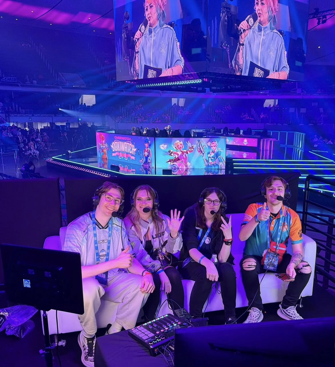 Cuatro personas con audífonos en un sofá, frente a un escenario de torneo