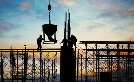 Trabalhadores da construção civil em silhueta sobre andaimes, içando um balde de concreto com guindaste ao entardecer, com o céu colorido ao fundo