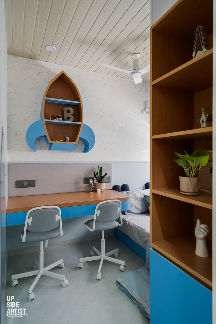 A dual study desk setup for children featuring a vibrant blue floating desk, grey chairs, and a custom rocket-shaped wooden wall shelf against starry wallpaper.
