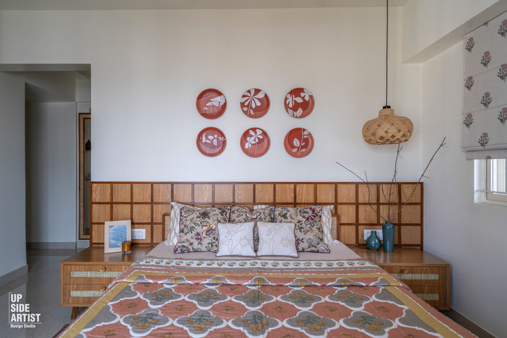 Earthy bohemian bedroom interior design showcasing a built-in light wood grid headboard, a bed with floral block-printed linens, terracotta wall art plates, and a wicker pendant light.