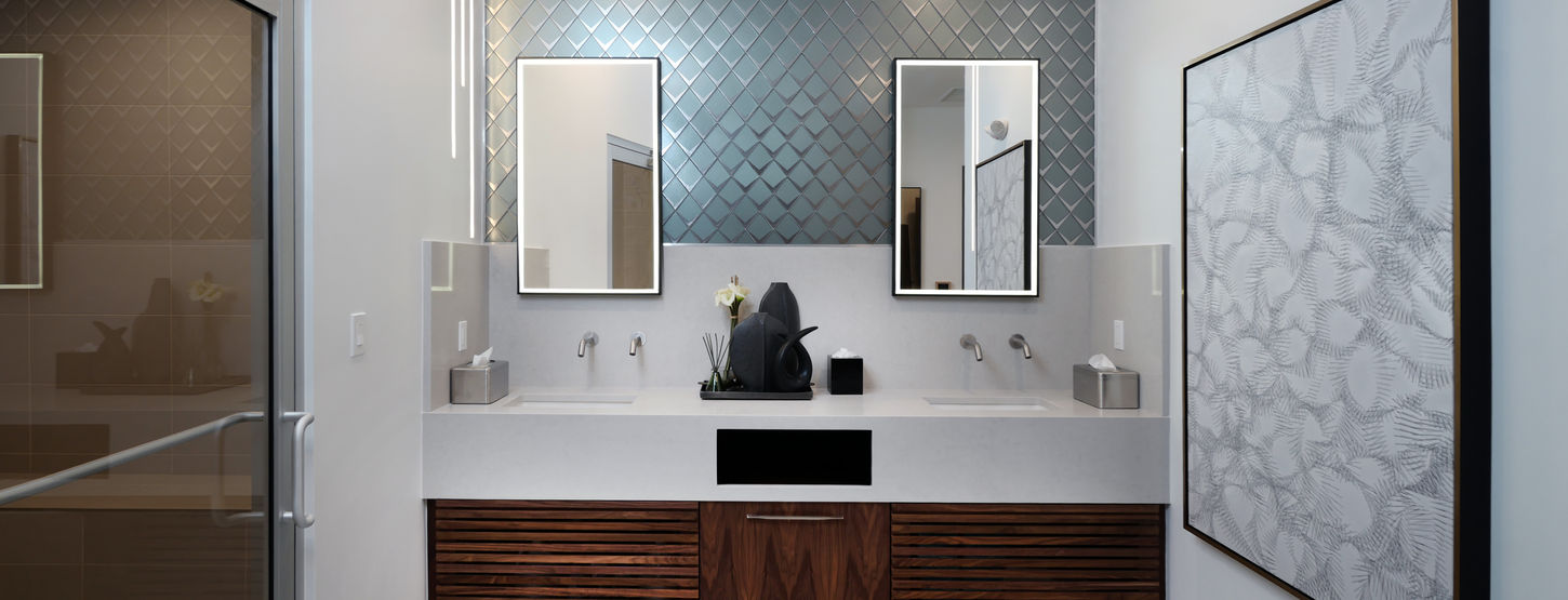 Women’s locker room vanity in Naples condo fitness center with dual sinks, geometric feature tile wall, illuminated mirrors, and custom wood cabinetry