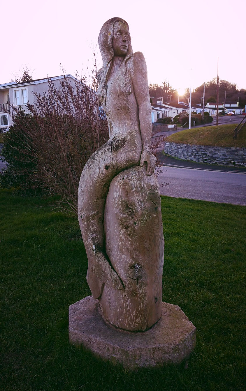 Mermaid sculpture on grassy lawn, against a pink sky backdrop with houses and trees in the background, evoking tranquility.