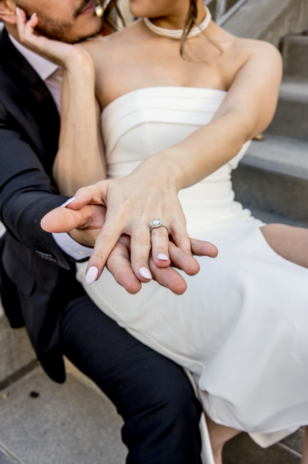 A couple sitting on steps, woman in white dress shows engagement ring. Man in suit gently holds her hand. Romantic and intimate mood.