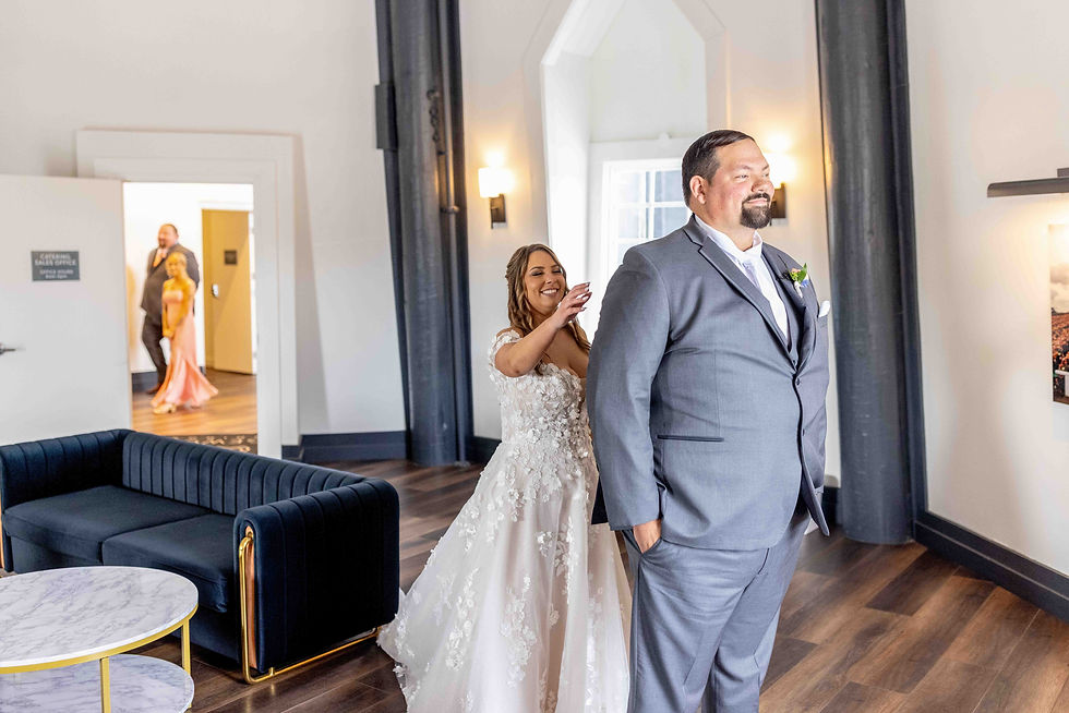 Bride in floral gown approaches groom in gray suit from behind, smiling in a bright room. Two people in the background. Cozy setting.
