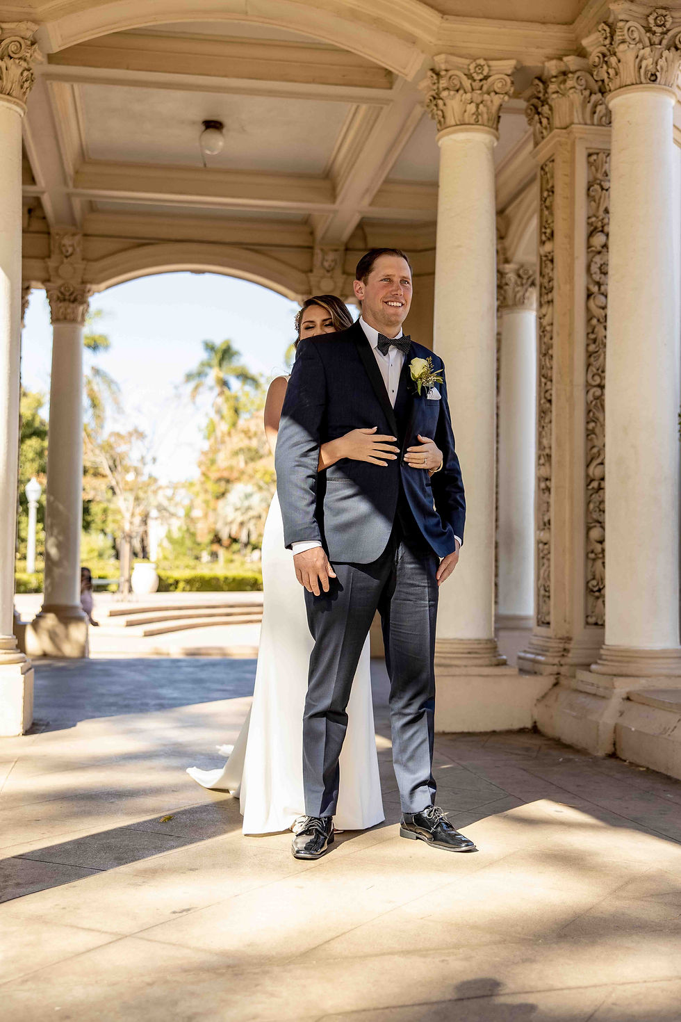 Bride hugs groom under ornate archway, both smiling. She wears white, he wears navy suit with yellow boutonniere. Sunlit outdoor setting.