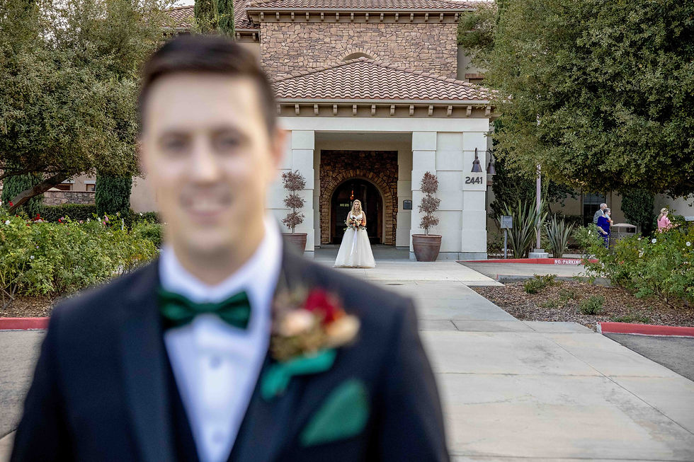 Blurred groom in suit with green bow tie in foreground, bride in gown holding flowers in background. Stone building with "2441" visible.
